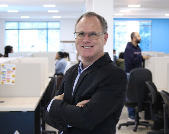 A imagem mostra Ademar Silva, CEO da Makro, sorrindo de braços cruzados, com mesas, cadeiras e alguns colaboradores da empresa Makro System ao fundo.