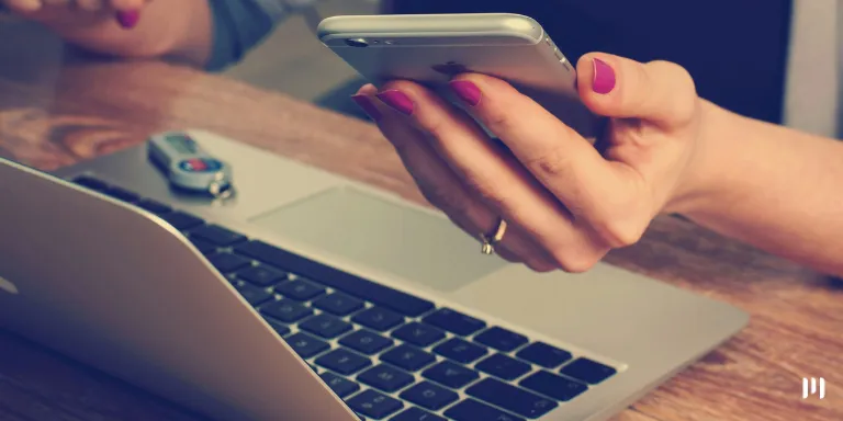 Mulher com esmalte rosa usa o computador para acessar o e-financeira. Ela também segura um celular na mão.