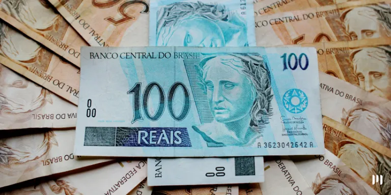 A imagem representa a moeda brasileira. No fundo várias notas de 50 reais formam um espiral e em cima delas duas notas de 100 reais formando o sinal de mais. A imagem representa a valorização do real. taxação de empreendedores informais