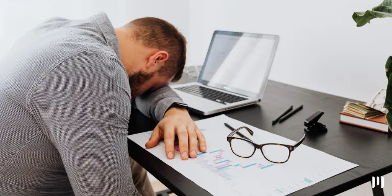 Homem deitado sobre o notebook, com óculos de grau e folhas de relatórios ao lado. A imagem representa a Síndrome de Burnout.