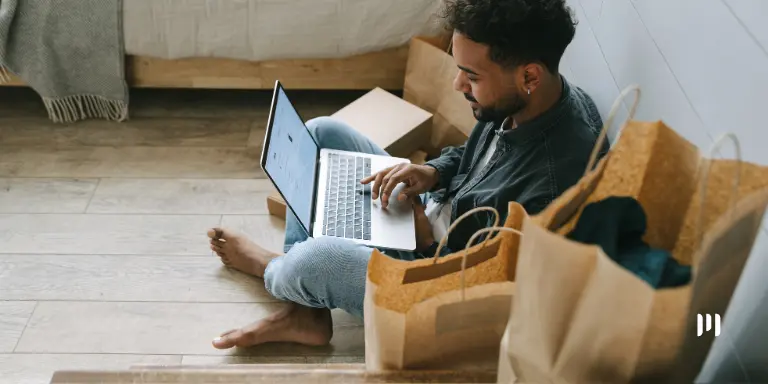 Homem usando laptop, cercado por sacolas de compras, na sala. Ele está vendo sobre a classificação tributária do IBS e CBS.